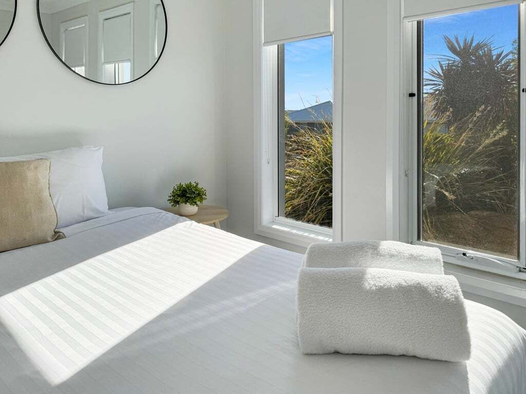 A bedroom with a bed, two pillows, a bedside table with a plant, and two windows with views of the outside.