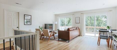 A spacious living room with a wooden floor, a brown sofa, a dining table with chairs, and a balcony with a view of trees.