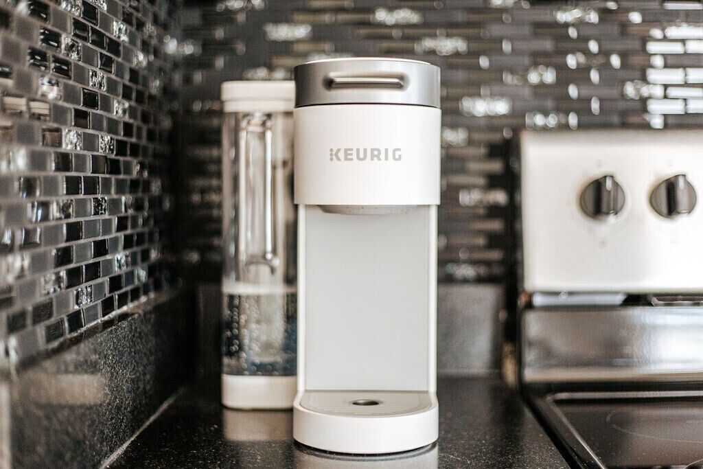 Une machine à café Keurig blanche est posée sur un comptoir de cuisine, avec un dosseret de carreaux noirs.