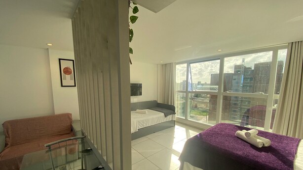 A modern hotel room with a glass partition, a bed with a purple bedspread, a sofa, and a large window offering a city view. - Bela Hospedagem - Wind Palace 606  (Cabedelo)