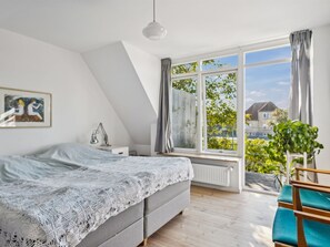 Ein Schlafzimmer mit Bett, Schreibtisch, Stuhl, Heizkörper und Fenster mit Blick auf Häuser und Bäume.