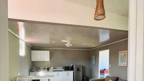Ett modernt kök med vita skåp, ett kylskåp och en mikrovågsugn. Det finns en matplats med ett vitt bord och två stolar. I vardagsrummet står en beige soffa med en gul kudde.