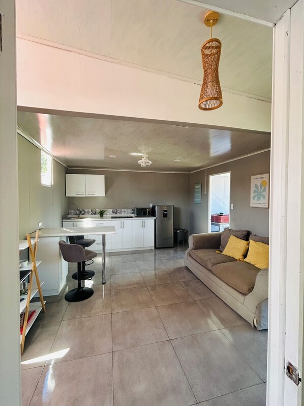 A modern kitchen with white cabinets, a refrigerator, and a microwave. There is a dining area with a white table and two chairs. A beige sofa is in the living area with a yellow cushion.