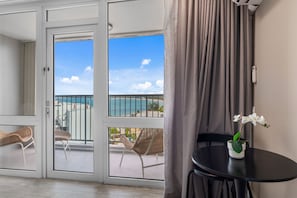 A room with a sliding glass door leading to a balcony with a view of the sea and beach. There is a small table with a potted orchid.