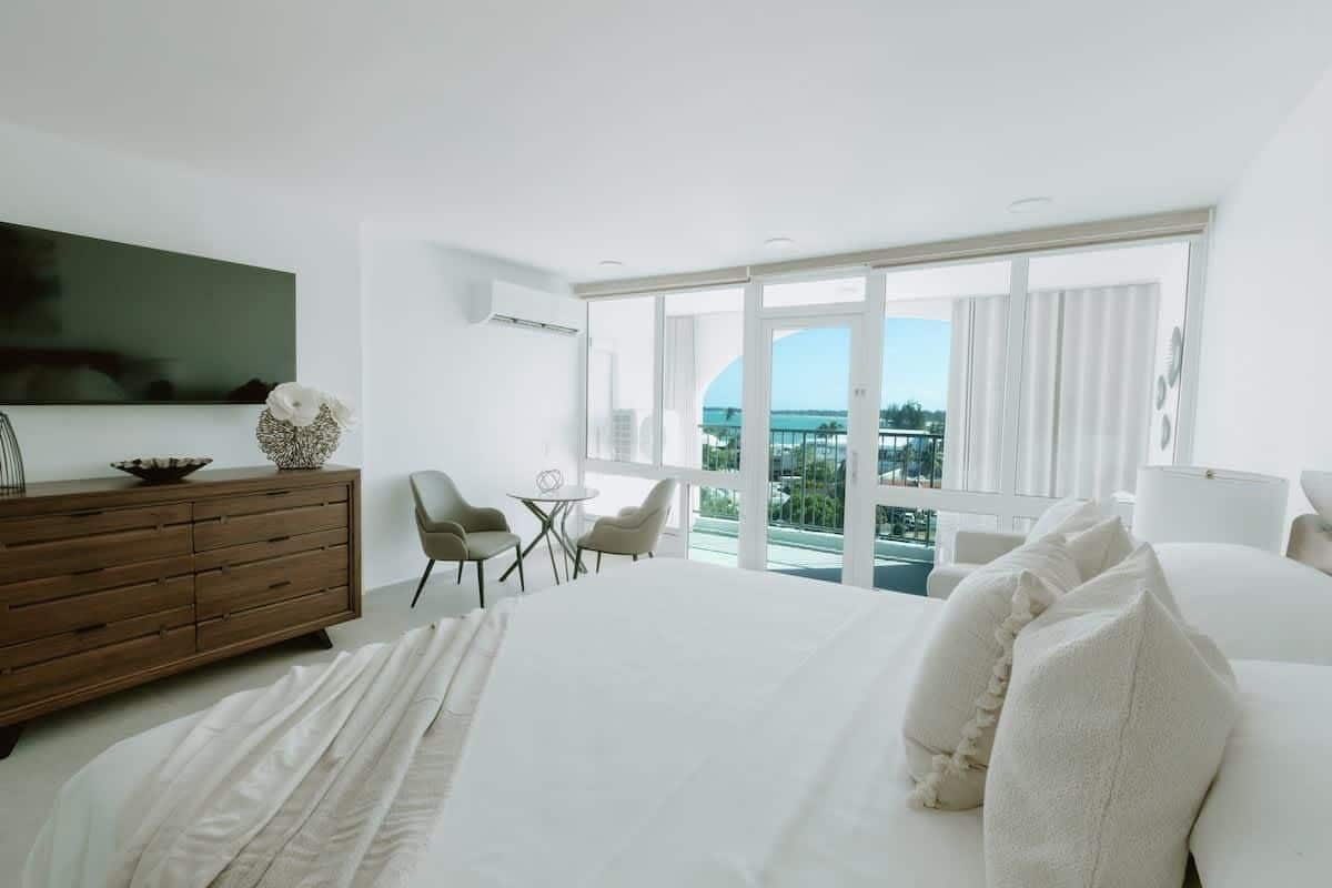 Una habitación de hotel moderna con una cama grande, un aparador de madera, un televisor montado en la pared, un balcón con vistas y una pequeña mesa con sillas.