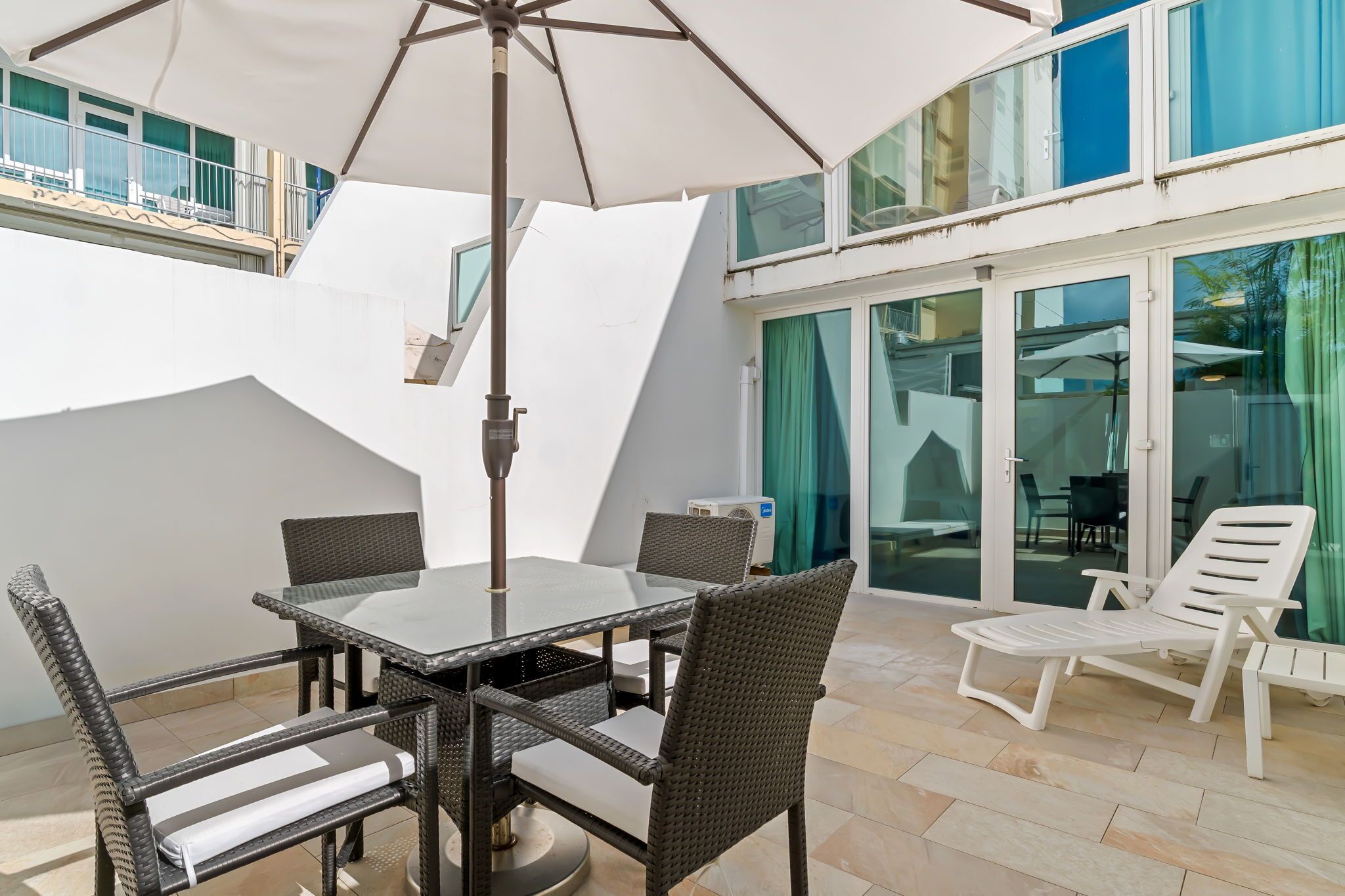 A patio with a glass table and chairs, a white umbrella, and a white lounge chair.