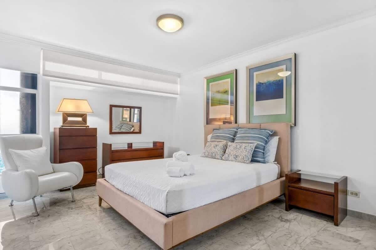 Un dormitorio moderno con una cama grande, una silla, una mesita de noche y dos cuadros en la pared.