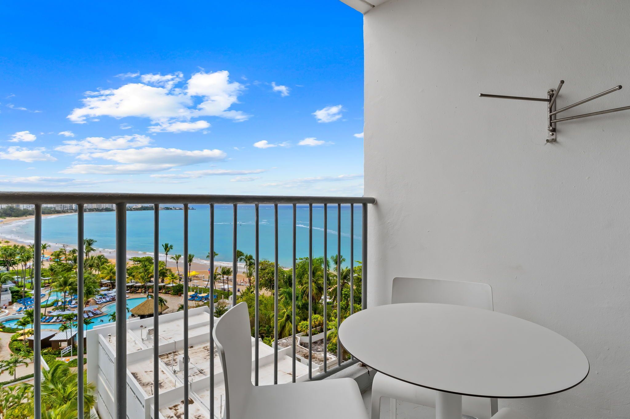 A balcony with a white table and chairs, overlooking a beach and pool area.