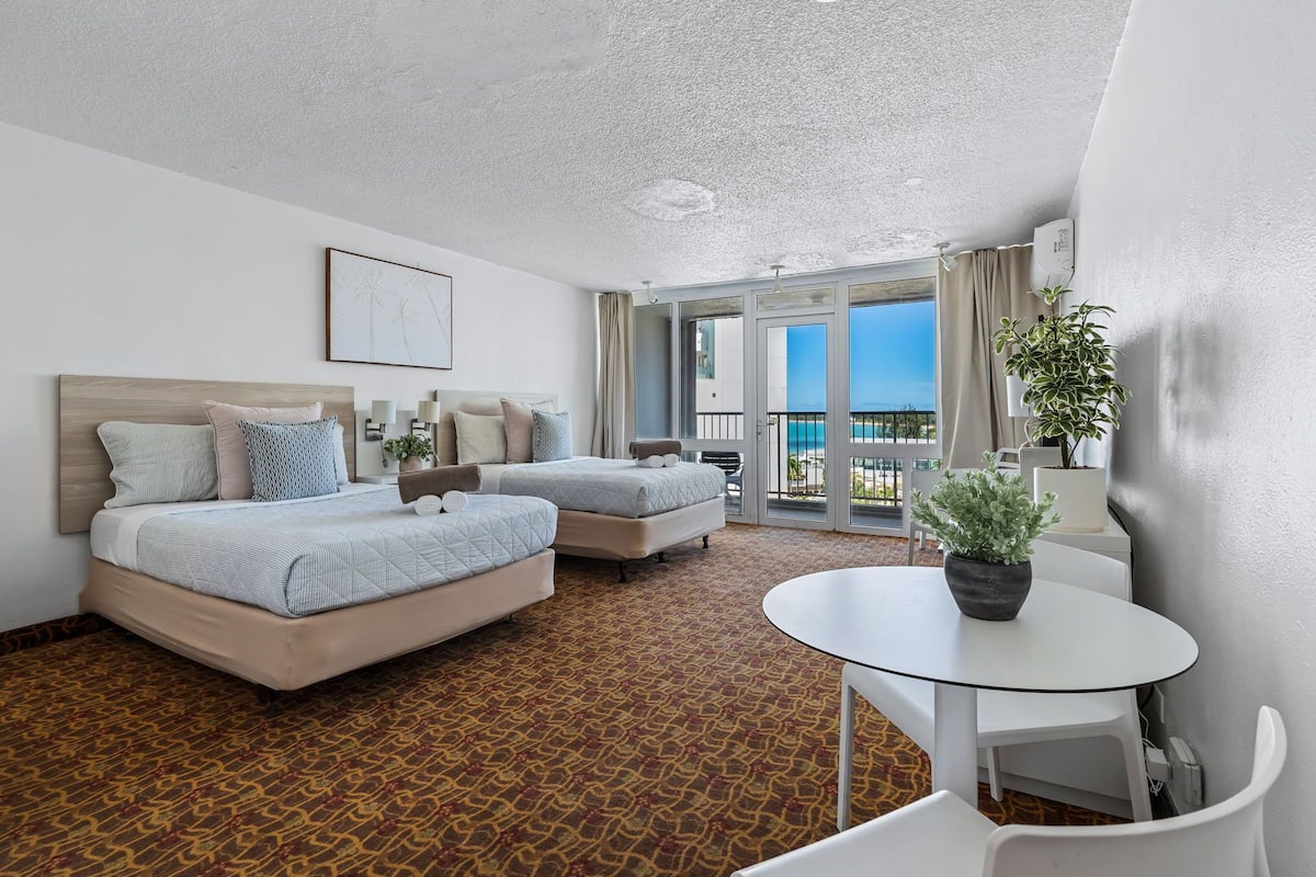 Habitación de hotel con dos camas, un balcón con vistas, una mesita y una planta en maceta.