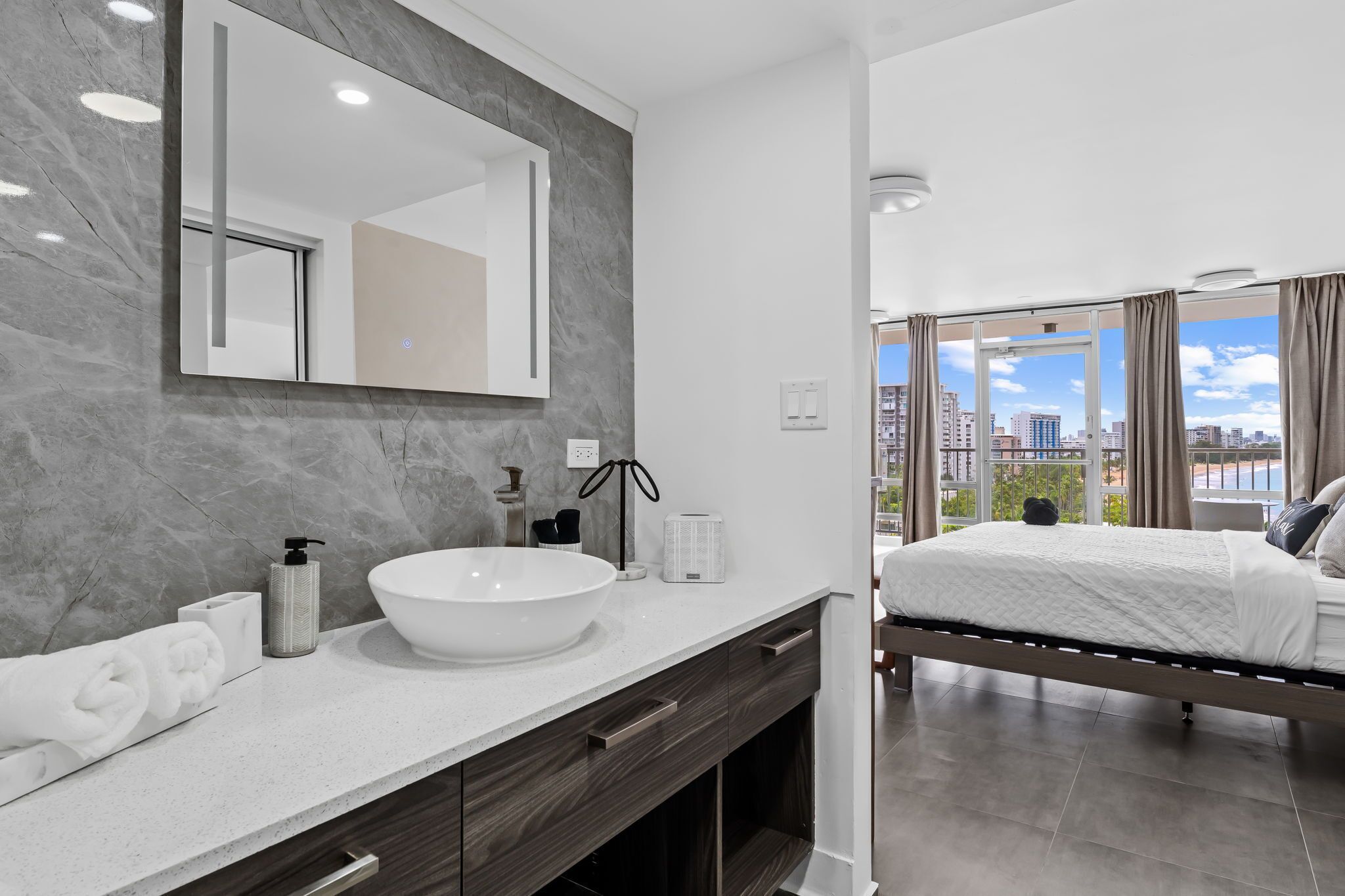 A modern bathroom with a large mirror, white sink, and a view of the city through large windows.