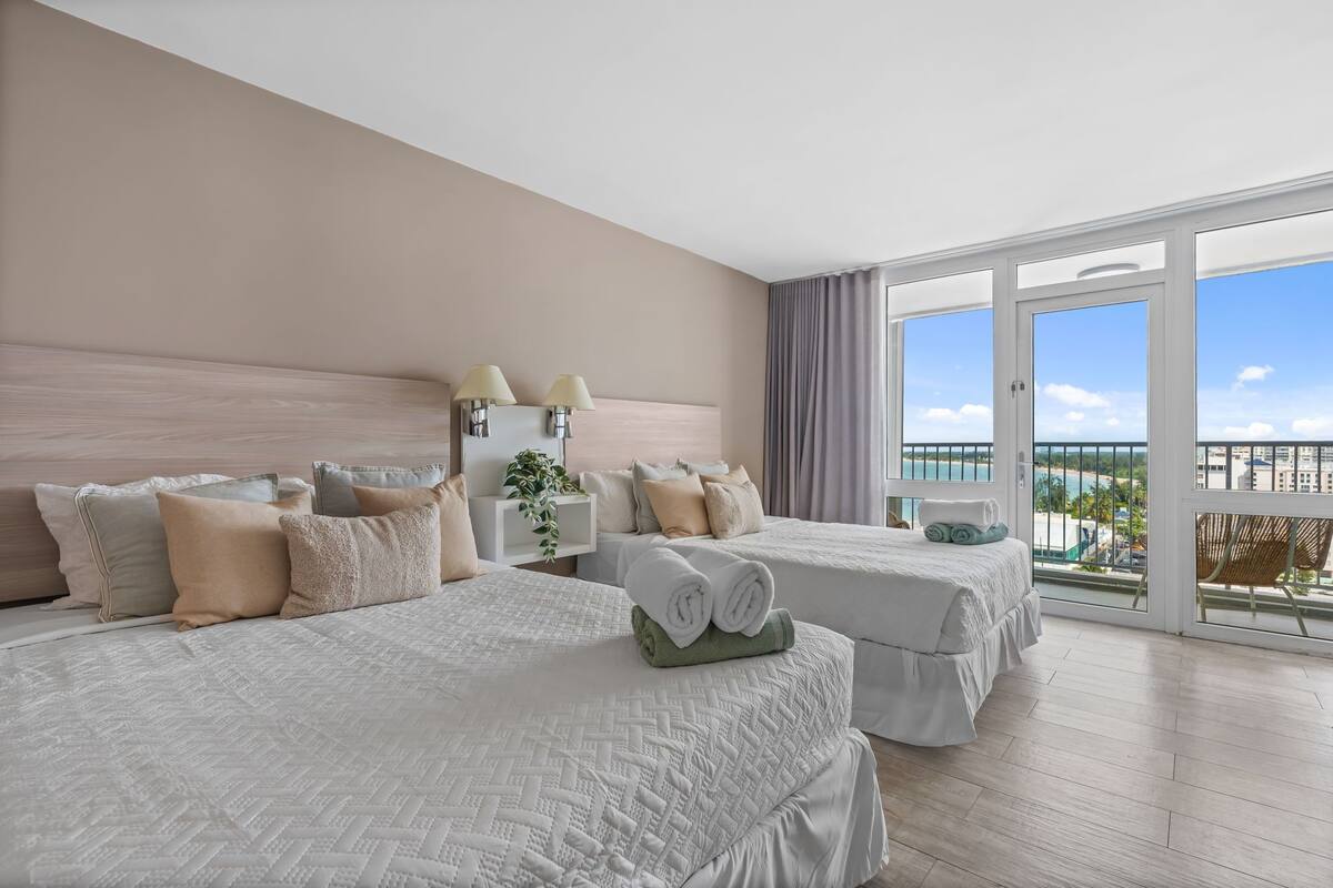 Habitación de hotel con dos camas, un ventanal con vistas, un balcón y una planta en la mesita de noche.
