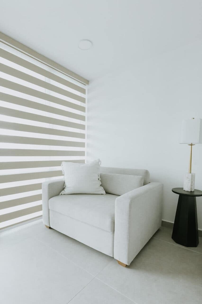 Un canapé blanc avec des coussins, une petite table d’appoint noire avec une lampe, et une fenêtre avec des stores horizontaux.