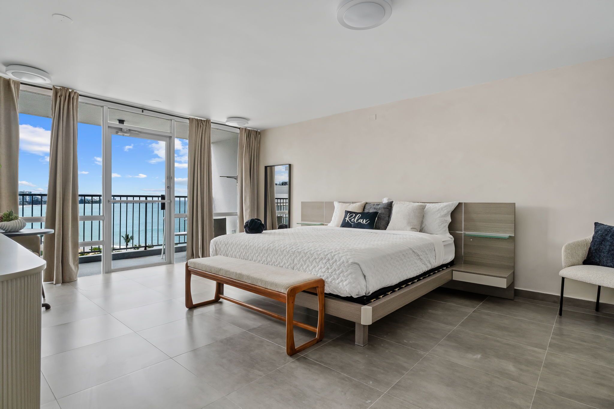 A modern bedroom with a large bed, a bench, a bedside table, a chair, and a view of the sea through large windows.