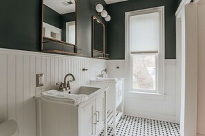 Une salle de bain avec un meuble-lavabo blanc, un grand miroir et une fenêtre avec des stores.