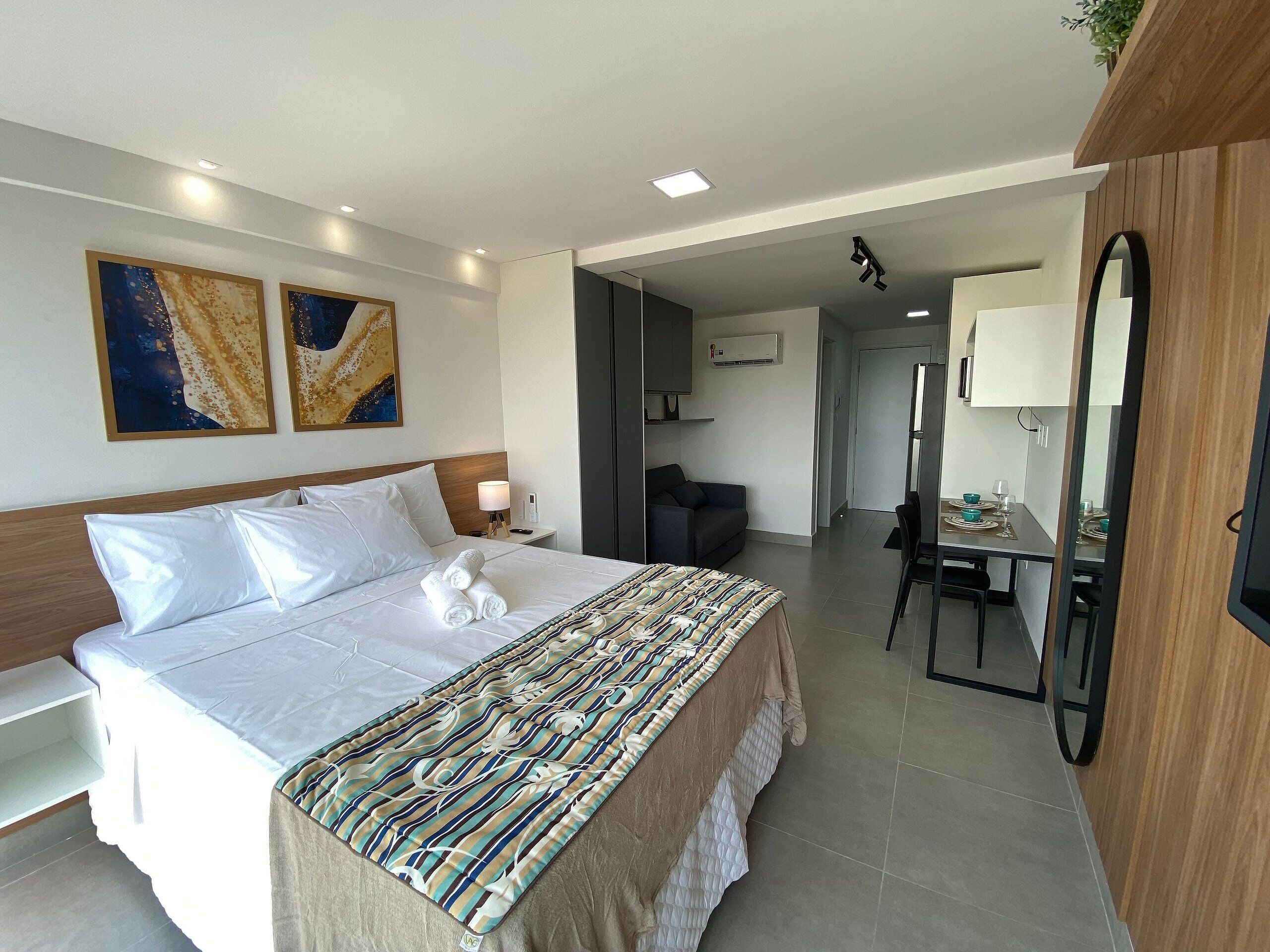 A hotel room with a bed, bedside table, lamp, artwork, and a view into the dining area.