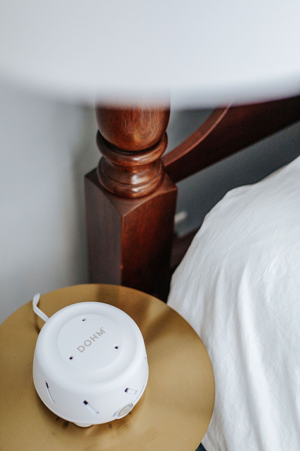 Un altavoz portátil blanco sobre una mesita de noche de madera y un almohadón blanco.