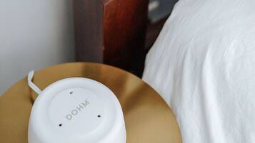 Un altavoz portátil blanco sobre una mesita de noche de madera y un almohadón blanco.