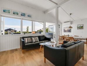 Ein großzügiger Wohnbereich mit einer schwarzen Ledersofa, einem Couchtisch und einem Essbereich mit Holzsesseln und Tisch. Große Fenster bieten Blick ins Freie.