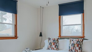 Ett sovrum med en säng, en takfläkt, två fönster med blå gardiner, en mönstrad matta och ett synligt eluttag.