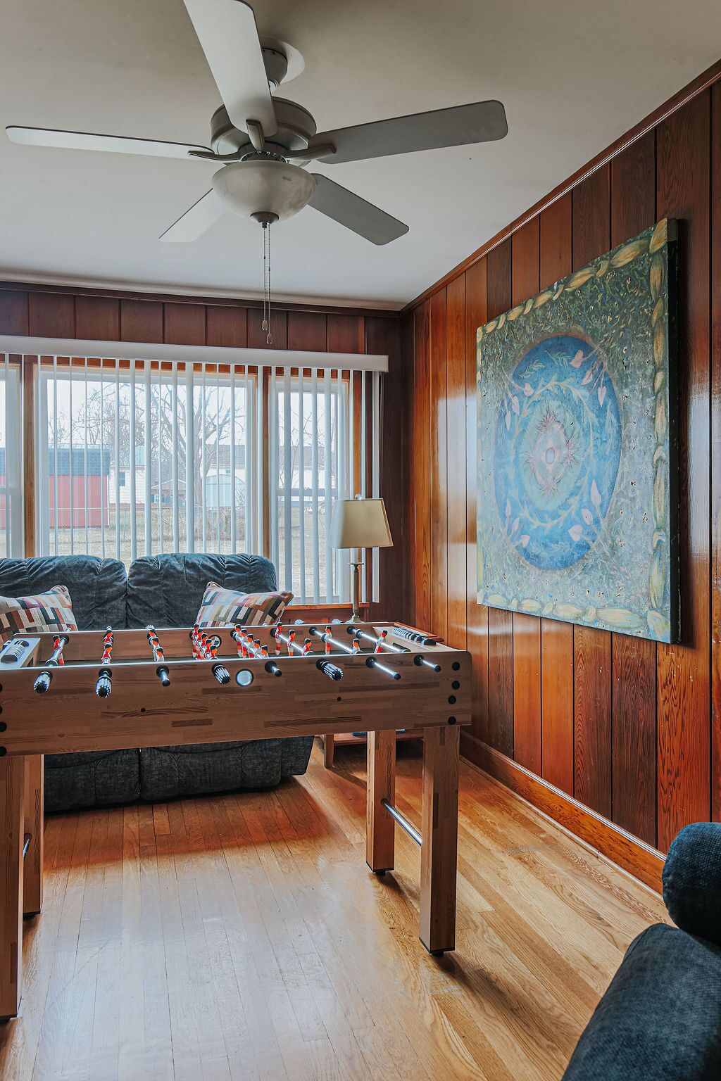 A room with a foosball table, a ceiling fan, a large painting, and a window with blinds.