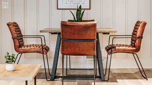 Una habitación con una mesa de madera, dos sillas de cuero, una planta en maceta y una obra de arte enmarcada en la pared.