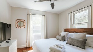 Un dormitorio con cama, almohadas, televisor, espejo y ventana.