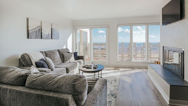A modern living room with a gray sectional sofa, a small coffee table, and a flat-screen TV mounted on the wall. Large windows provide a view of the cityscape. - Mountain View Vista (Nellysford)
