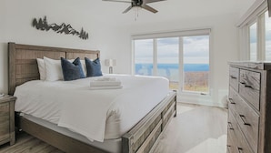 A bedroom with a large bed, a ceiling fan, a nightstand, a dresser, and a window with a view of the sky and landscape. - Mountain View Vista (Nellysford)