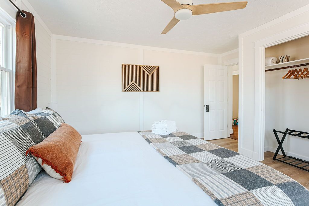 A bedroom with a bed, a ceiling fan, a geometric wall decoration, a folded towel on the floor, and a closet with hanging clothes.