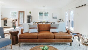 Uma sala de estar moderna com um sofá de couro marrom, uma mesa de centro redonda de madeira e uma área de jantar com mesa e cadeiras brancas.