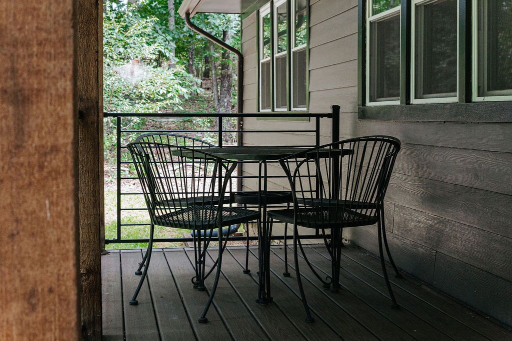 A covered outdoor seating area with metal chairs and a table, surrounded by a wooden deck and a house with white siding and green window frames.