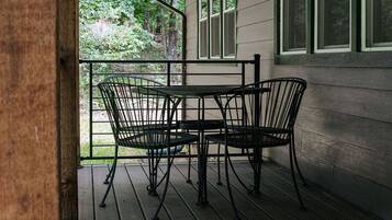 A covered outdoor seating area with metal chairs and a table, surrounded by a wooden deck and a house with white siding and green window frames.