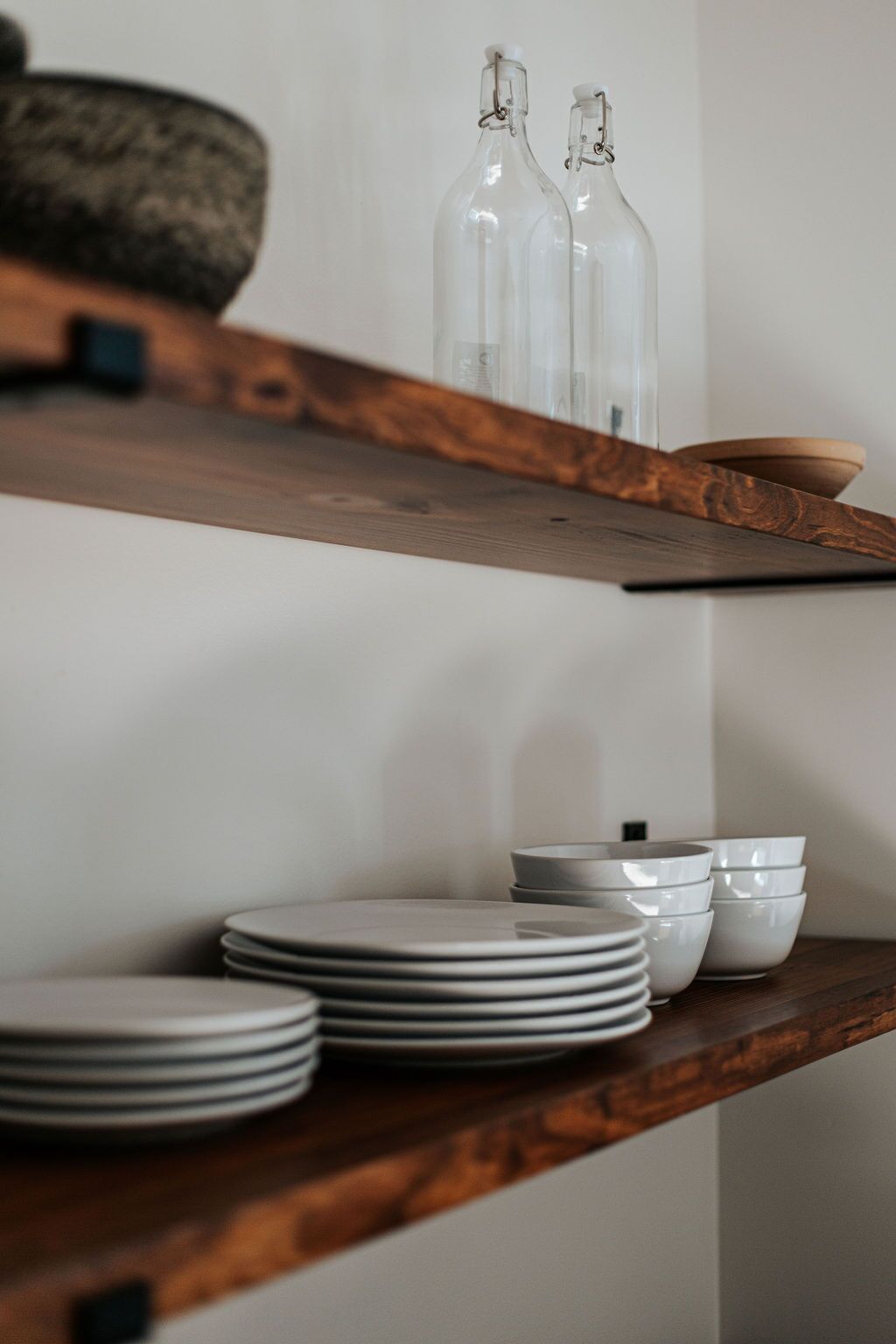 A wooden shelf with stacked white plates and two clear glass bottles.