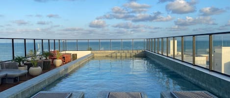 Piscina infinita no terraço com espreguiçadeiras e plantas em vasos, com vista para o oceano.