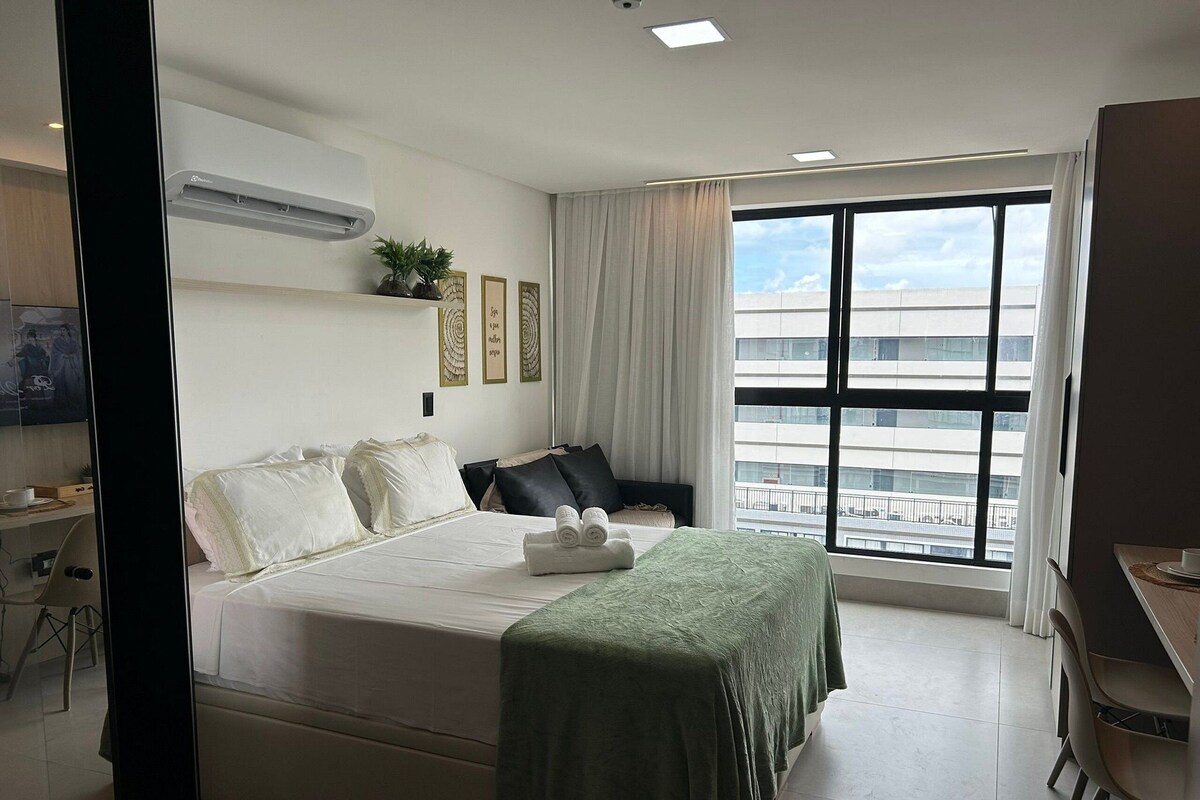 Quarto de hotel com cama, escrivaninha, cadeira, janela e vista para edifícios, e um aparelho de ar condicionado.
