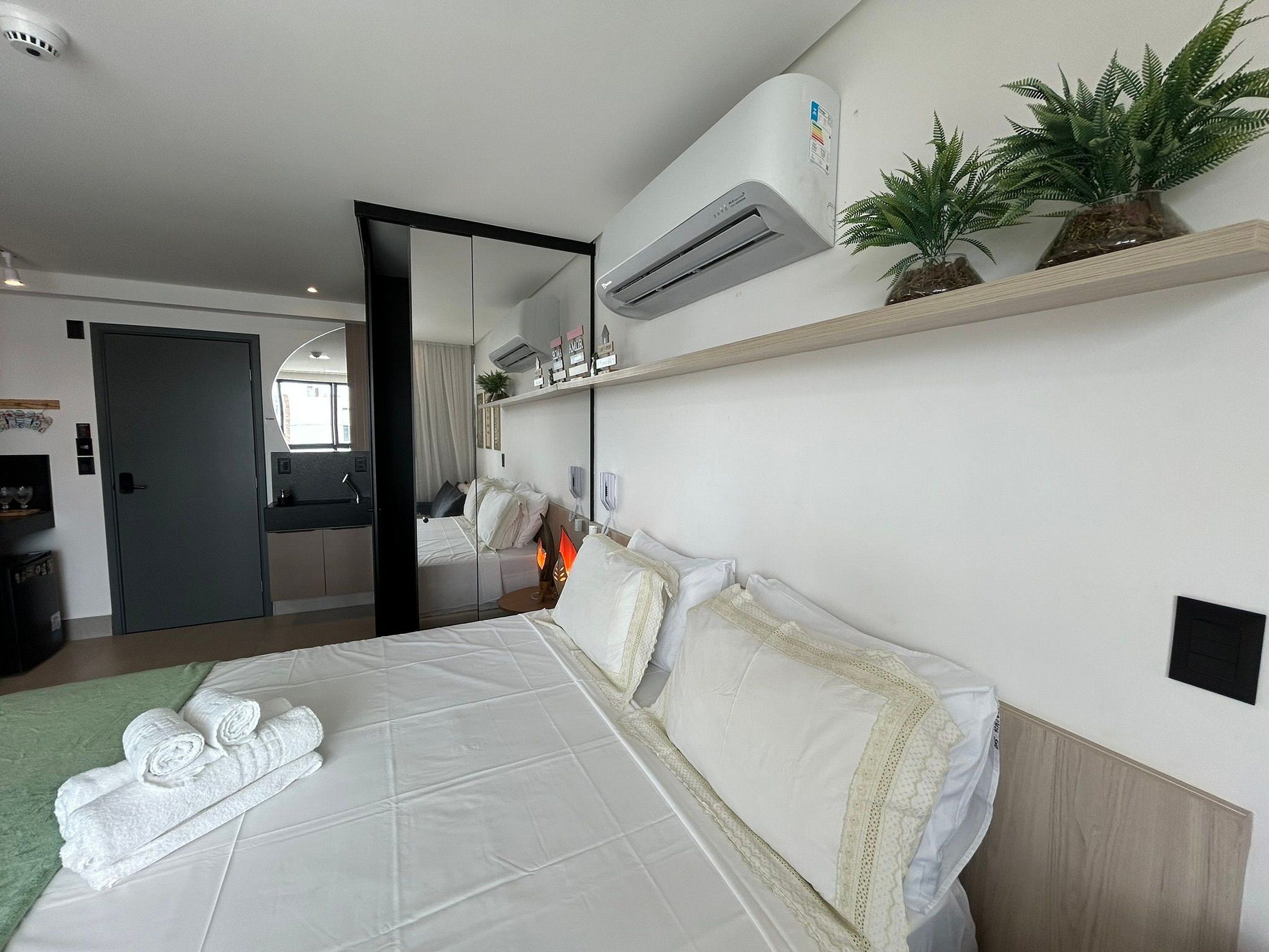 Una camera d'albergo con un letto, uno specchio, un condizionatore, uno scaffale con oggetti decorativi e una porta.