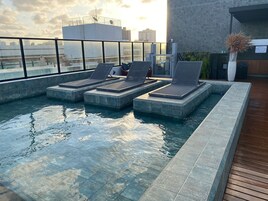 Una piscina en la azotea con sillones de descanso, una terraza de madera y vistas a la ciudad.
