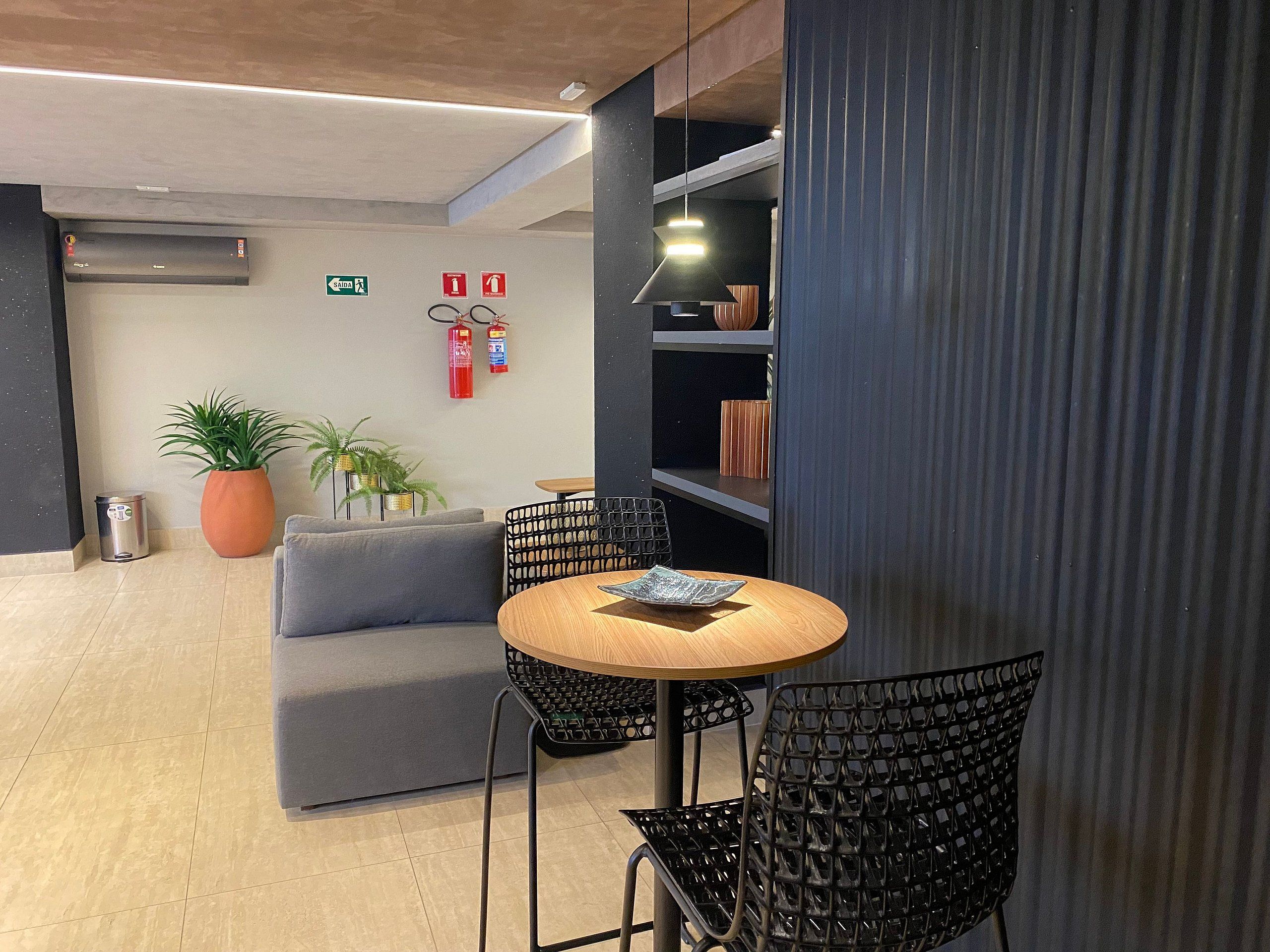 Un intérieur moderne avec une table ronde en bois, des chaises noires en résille, un canapé gris, des plantes en pot et des extincteurs fixés au mur.