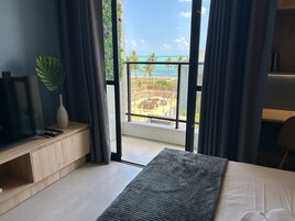 Quarto com uma janela grande, cama, televisão, planta e vista para uma praia e um canteiro de obras.