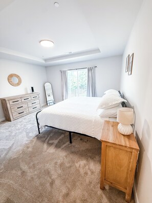 A bedroom with a bed, a wooden nightstand, a dresser, a mirror, and a window with curtains.