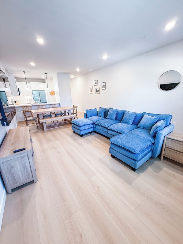 A modern living room with a sectional sofa, wooden flooring, a dining area with a table and chairs, and a kitchen in the background.