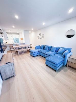 Una sala de estar moderna con un sofá seccional, piso de madera, una zona de comedor con mesa y sillas, y una cocina al fondo.