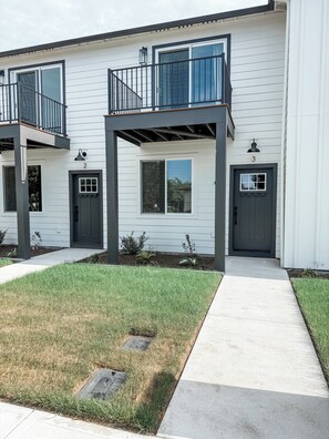 A two-story residential building with a covered front porch, a small front yard, and a visible manhole cover.