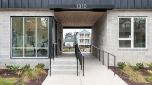 En moderne bygning med tallet '1310' over indgangen, store vinduer, en trappe med gelænder og et lille haveområde.