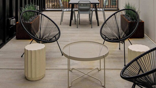 En moderne udendørs terrasse med bord og stole, lyskæder og udsigt til bygninger i baggrunden.