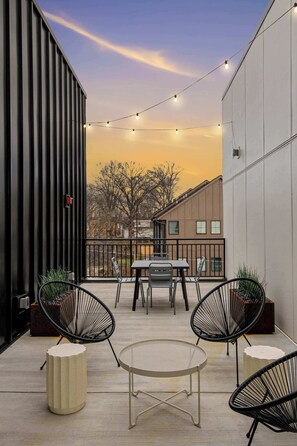En moderne udendørs terrasse med bord og stole, lyskæder og udsigt til bygninger i baggrunden.