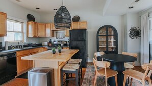 A modern kitchen with wooden cabinets, a central island, a black refrigerator, and a dining area with a round table and chairs. - Oasis on 5th (Charlottesville)