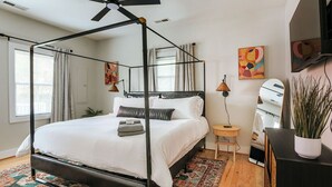 A four-poster bed with a canopy, a small bedside table with a lamp, and a potted plant on the right. - Oasis on 5th (Charlottesville)
