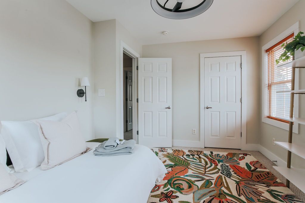 Un dormitorio con cama, dos puertas blancas, una ventana con persianas y una lámpara en el techo.