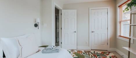 Un dormitorio con cama, dos puertas blancas, una ventana con persianas y una lámpara en el techo.